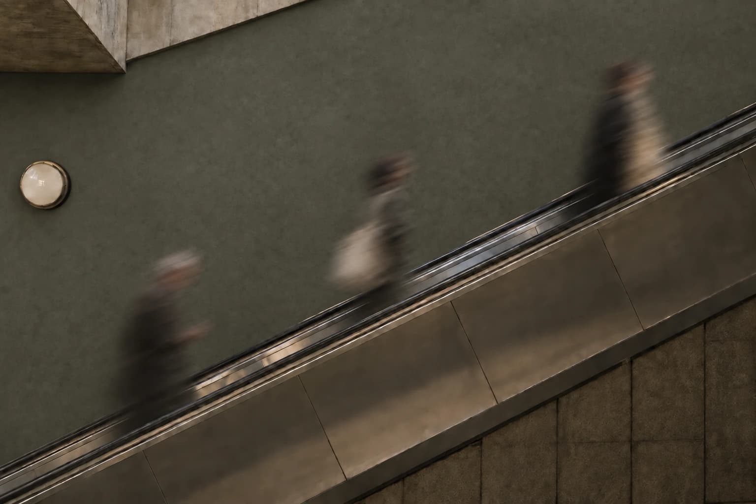 Three abstract commuters blurred by long exposure on an escalator beside a muted olive-gray wall.